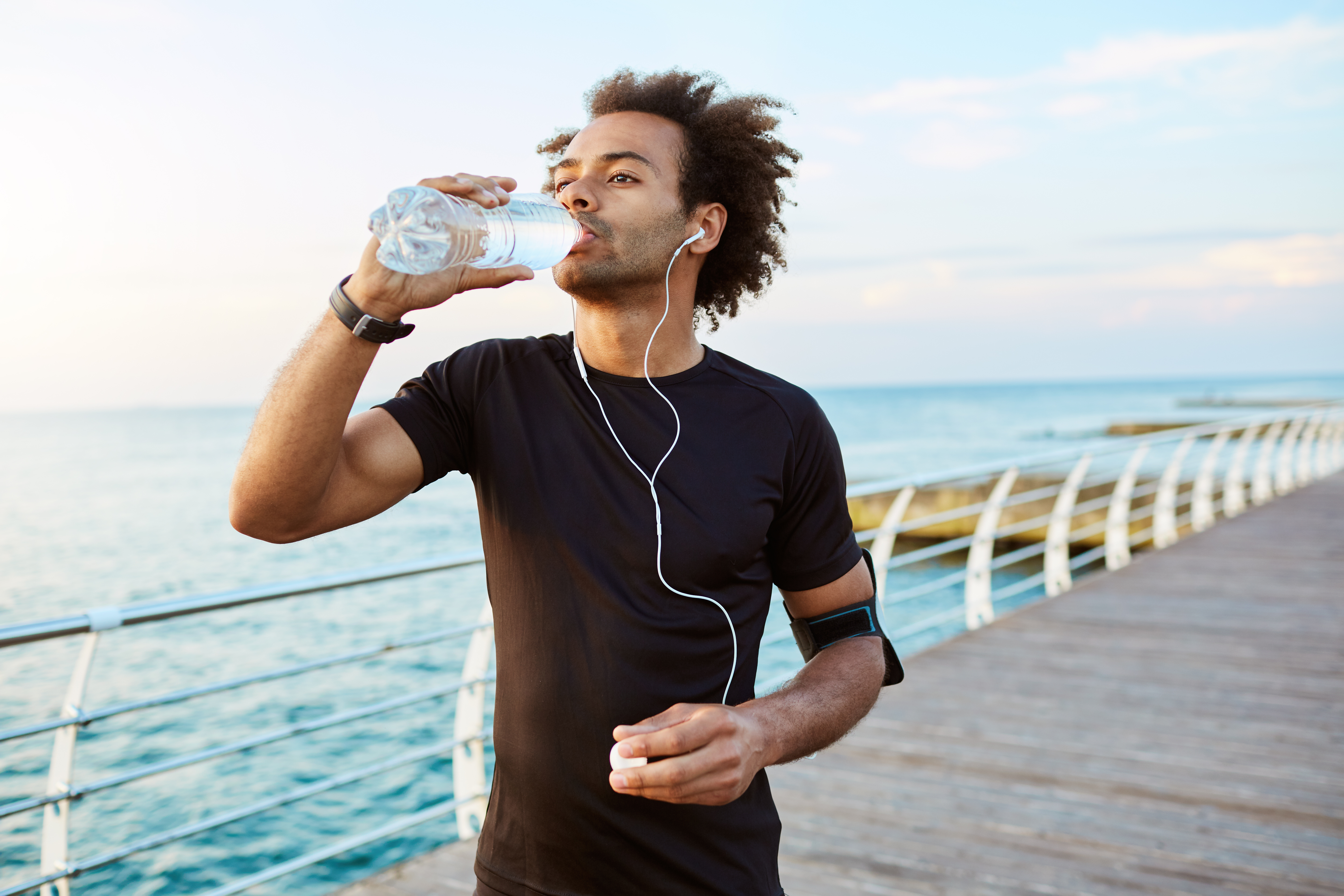 Homem se exercitando faz uma pausa para beber água, usando fones de ouvido e roupa esportiva, em um calçadão à beira-mar ao pôr do sol.