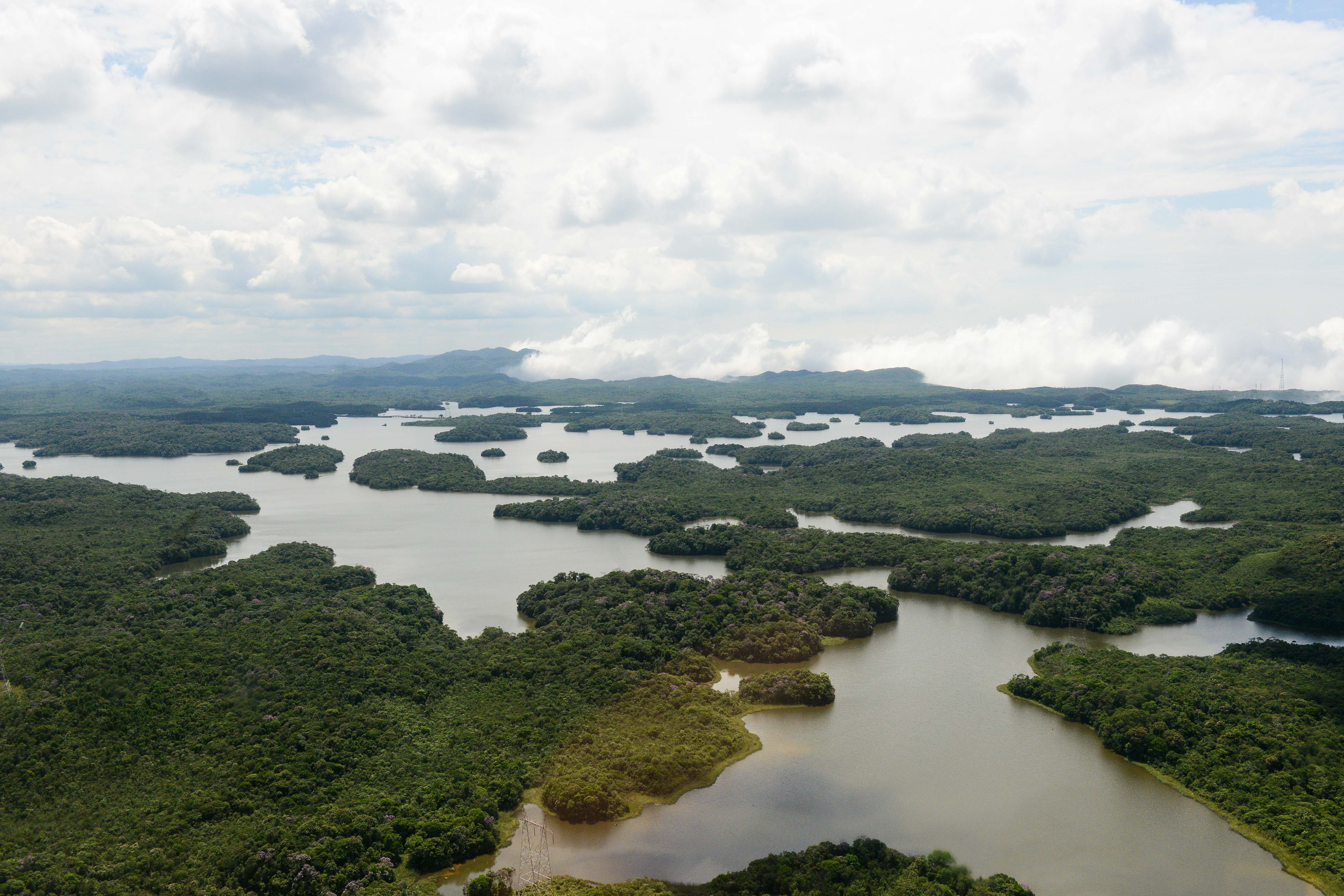Uma vasta paisagem aérea com partes sinuosas da represa Billings entre grandes áreas de floresta densa. As ilhas verdes aparecem espalhadas pela água calma, sob um céu nublado e claro.
