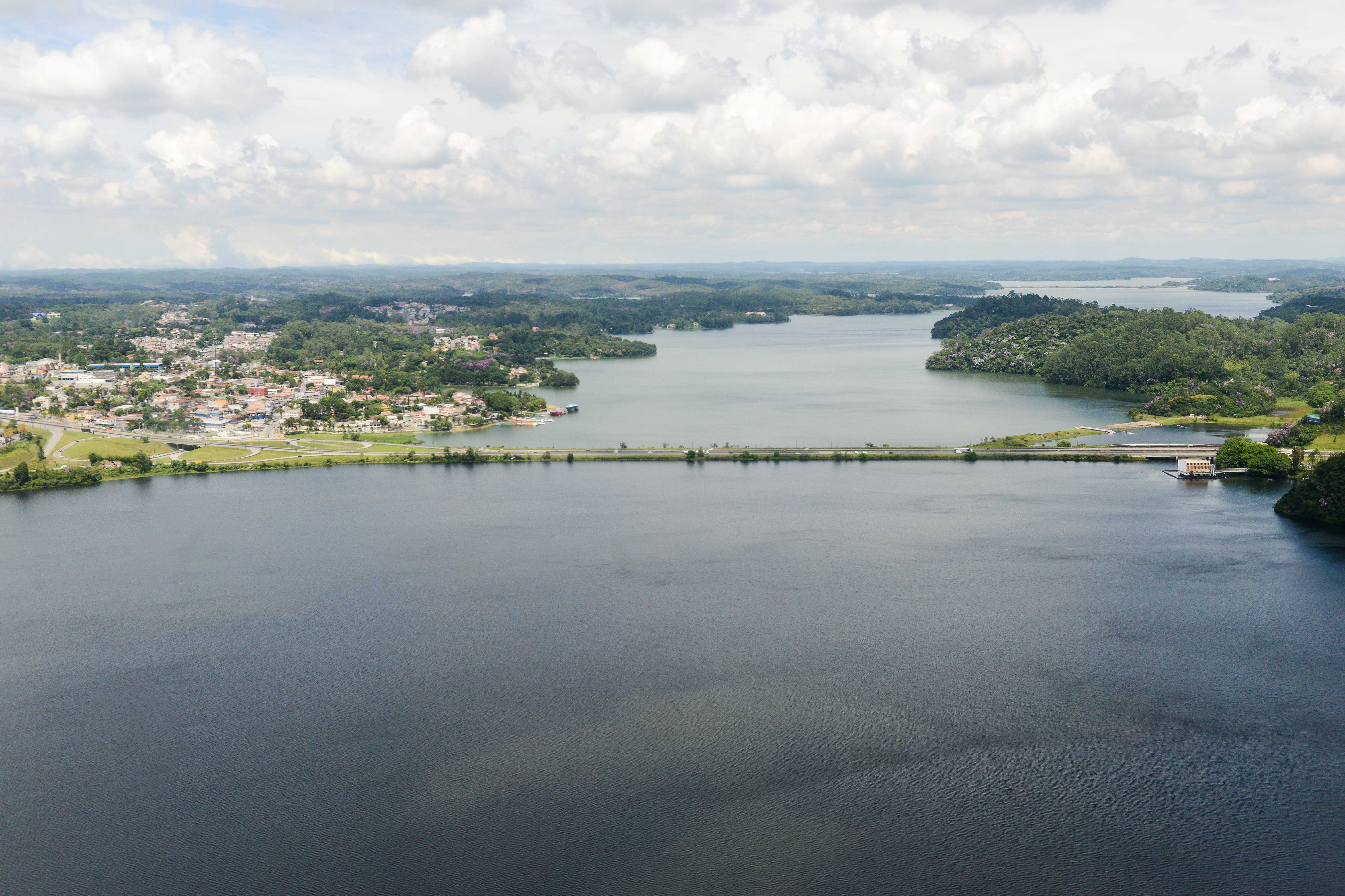 Vista aérea da Represa Billings: um grande espelho d’água escuro cercado por áreas verdes e uma cidade ao fundo. Há uma longa barragem ligando as margens, e o céu nublado reflete suavemente na superfície tranquila do reservatório.