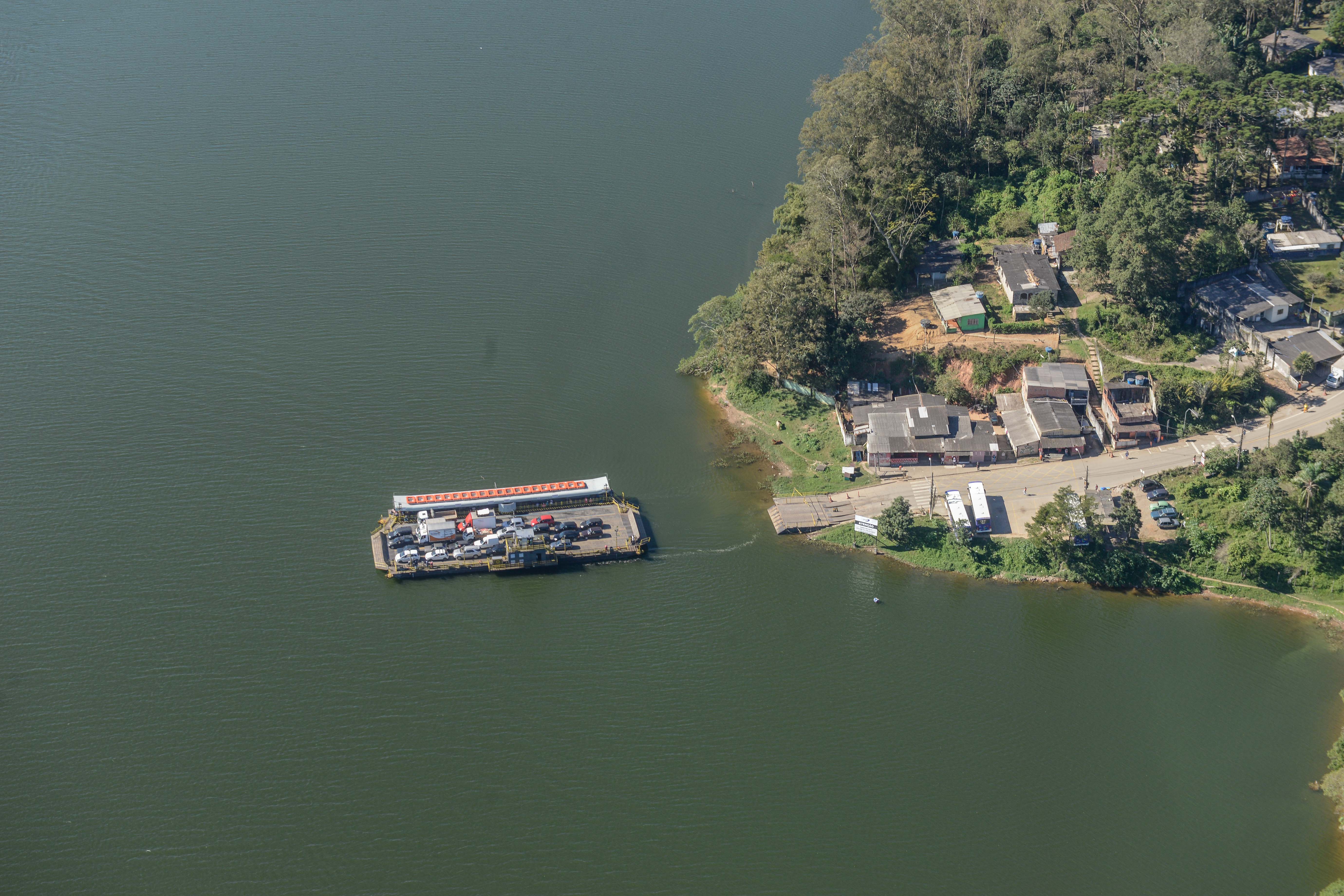 Uma balsa cruza a Represa Billings levando carros e motos. Ela está próxima de uma pequena área urbana à beira d’água, com casas simples e muitas árvores ao redor. A água é calma e verde-escura, contrastando com o verde da vegetação próxima.